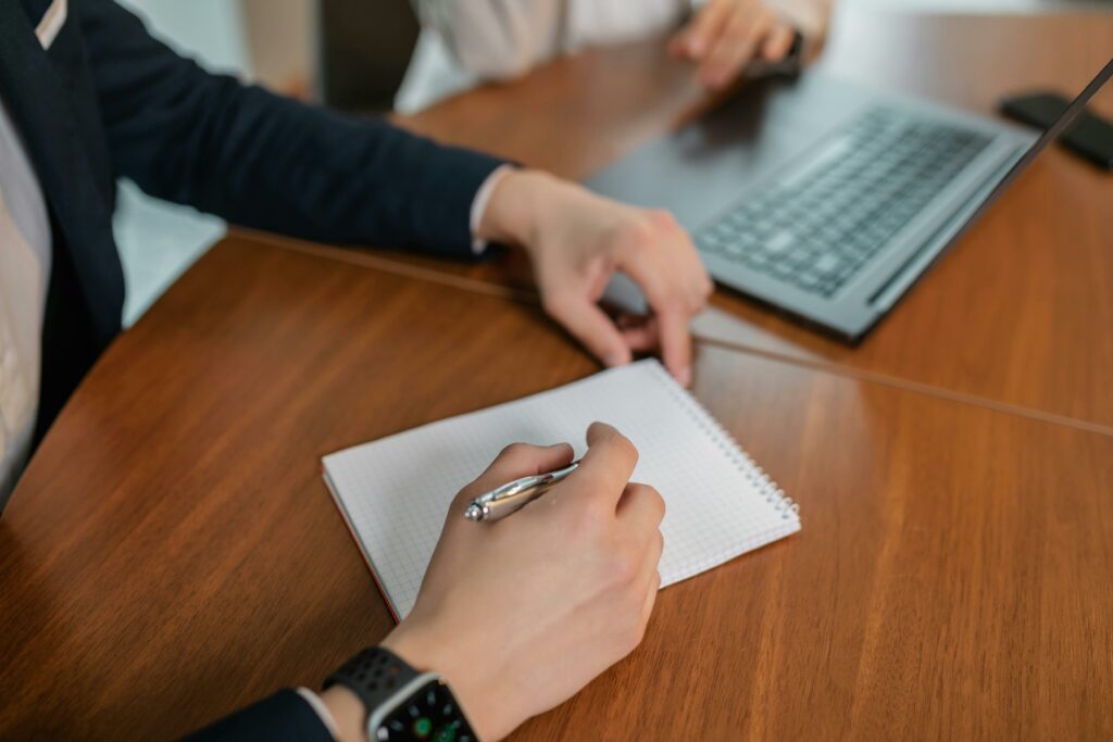 pexels-photo-8133853-8133853 Close-up of hands writing notes on paper during a business meeting indoors with a laptop on a wooden table.